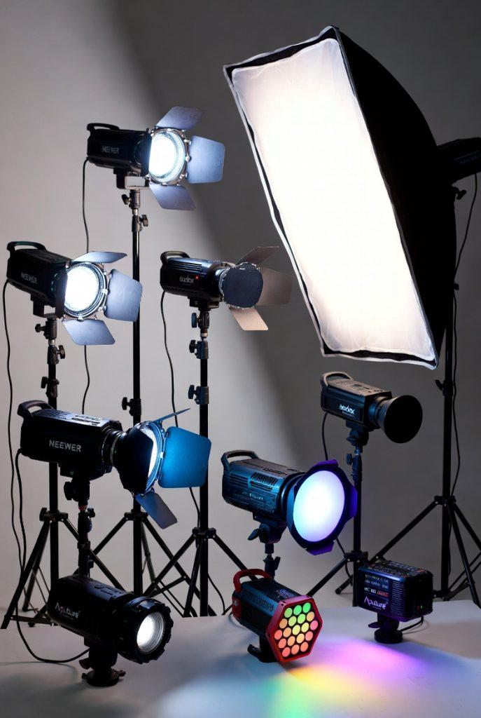 Assortment of budget-friendly and versatile LED lights for documentary filmmaking displayed on light stands: Aputure MC RGBWW pocket light with colorful hexagonal output, Neewer bi-color panel with barn doors, Godox SL series fresnel-style spotlight, large softbox with diffusion, and smaller RGB accent lights, all set up against a neutral gray backdrop to demonstrate practical lighting options for interviews and run-and-gun shoots.