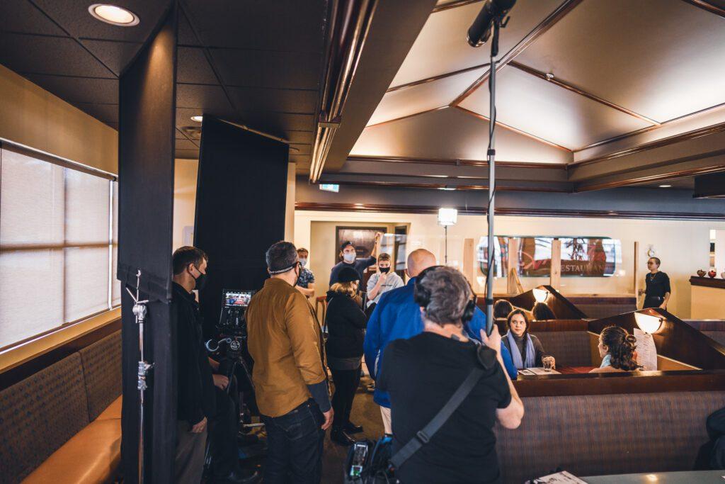 Scene from 'Going Home': Actors and crew in a restaurant, with camera and filmmaking equipment.