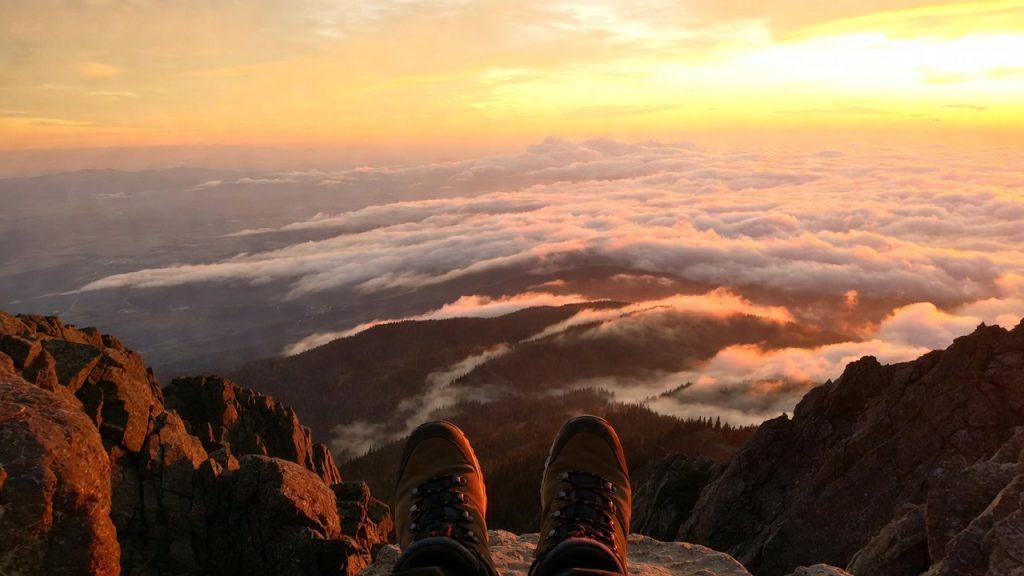 First-person view of a hiker's sturdy brown hiking boots resting on the rocky edge of a high mountain summit, looking out over a vast sea of fluffy white clouds below at sunrise. Warm golden-orange light from the rising sun bathes the scene, illuminating the distant mountain peaks and creating a peaceful, inspiring atmosphere of achievement, freedom, and the triumph of reaching a goal after a challenging climb.