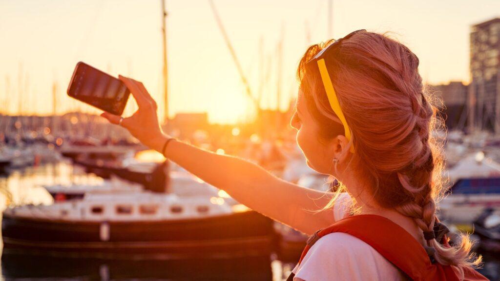 Smiling woman holding a smartphone, capturing the vibrant hues of a stunning sunset with her camera.