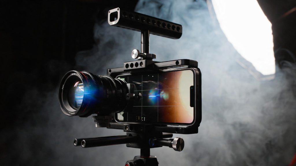 Cinematic product photography of professional filmmaker's smartphone rig in 2026. Phone mounted with T-Series anamorphic lens, variable ND filter, follow focus attachment. Screen displaying perfectly exposed cinematic footage with authentic blue lens flares. Dark moody studio lighting with dramatic rim lights, shallow depth of field focusing on lens elements, smoke machine adding atmosphere, professional cinematography gear aesthetic, shot on RED, 8k detail, commercial filmmaking style