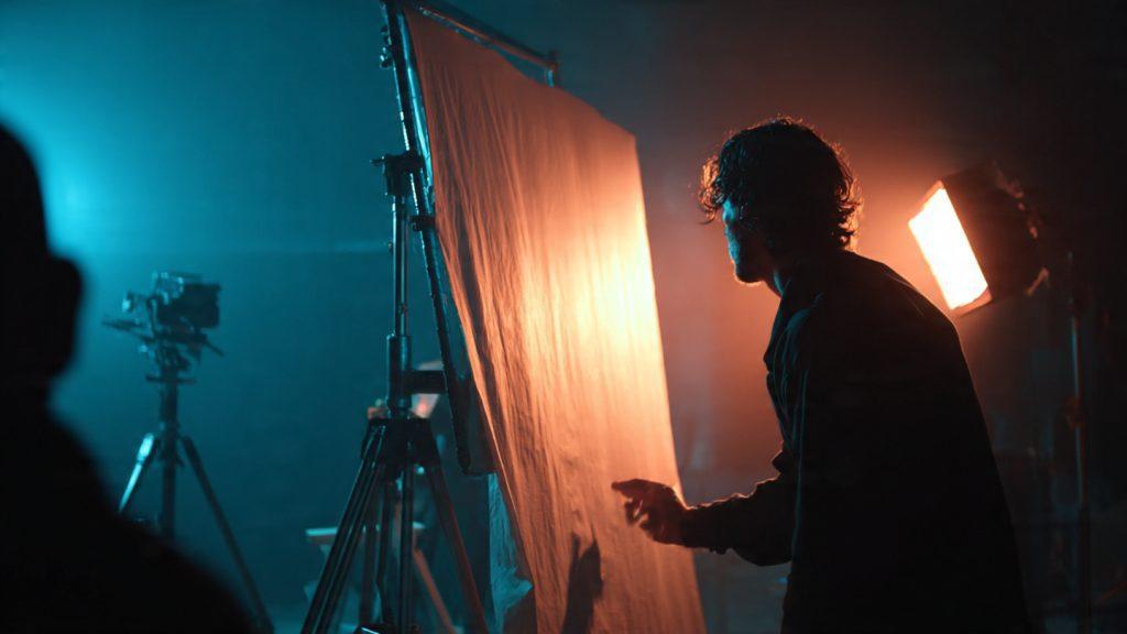 create an image based on this prompt"Filmmaker adjusting a V-shaped book light rig on a dark indie film set, with unbleached muslin fabric stretched across a 4×4 frame, visible C-stand hardware, and soft wraparound light hitting an actor's face—shot in moody blue/orange color grading"