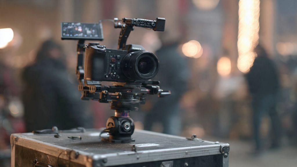 Close-up of a DJI RS 3 gimbal with motors locked in position, Sony a7 IV mounted, sitting on a director's monitor case between takes on set, crew activity blurred in background, photorealistic