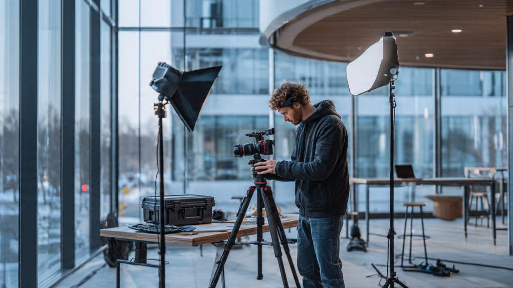 Filmmaker setting up a Sony a7 IV on a DJI RS 3 Pro gimbal at a corporate client location — modern glass office building, Aputure 300d II on a light stand with softbox, DJI Air 3 drone case open on a table in background, professional but not staged, photorealistic