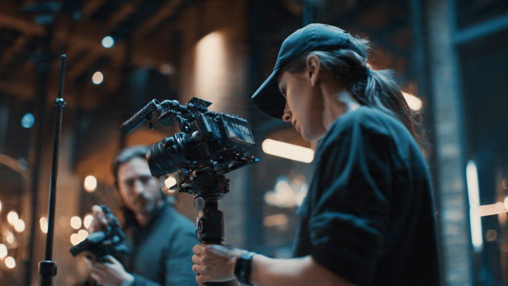 Small two-person film crew on a narrative short set: one person operating a DJI RS 3 gimbal with a mirrorless camera, another holding a DJI Pocket 3 for a secondary angle, Aputure Amaran 200d on a stand in background, interior location with practicals visible, photorealistic