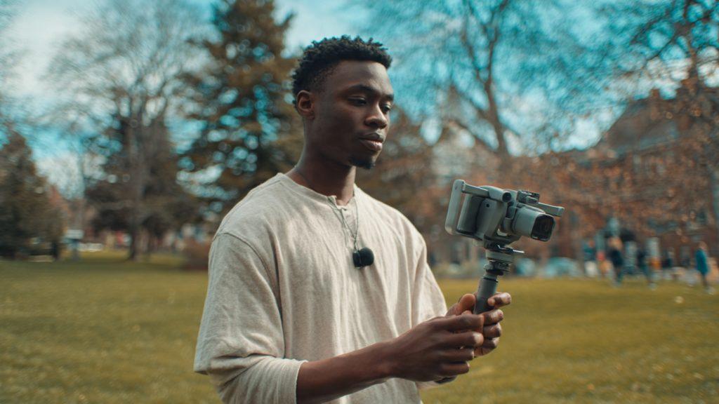First-time filmmaker in a park practicing with a DJI OM 6 and smartphone, slightly awkward stance, clearly learning, small DJI Mic Mini clipped to shirt, afternoon light, nobody watching, photorealistic
