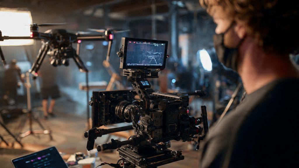 Single filmmaker operating a DJI Ronin 4D on a high-end commercial set, visible LiDAR on camera, Mavic 3 Pro Cine on a landing pad nearby, Aputure 600d Pro with large softbox in background, client watching a monitor off to the side, photorealistic