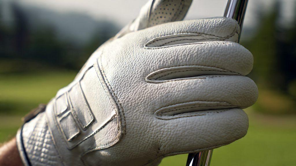 A hand wearing a white leather golf glove with visible stitching holds the grip of a golf club, with a blurred green fairway in the background