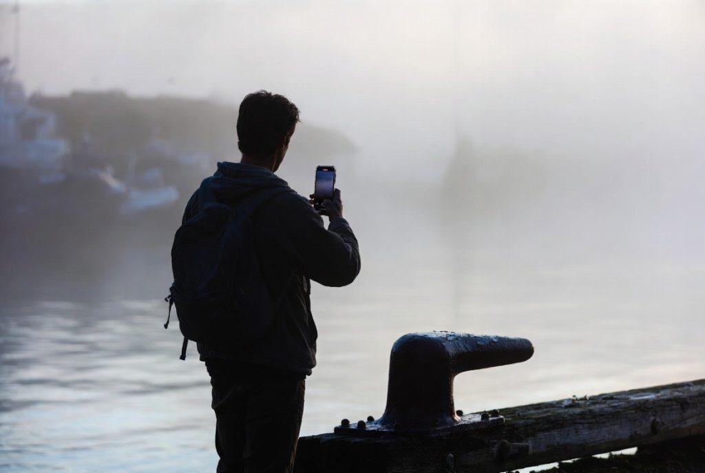 Solo filmmaker in low fog at Victoria BC inner harbour, 5:45 AM, leaning a smartphone against a weathered dockside bollard, minimal gear, realistic documentary atmosphere, no artificial lighting
