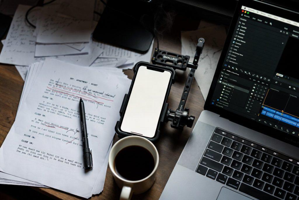 Prompt: "A top-down 'flat lay' of a filmmaker's desk in 2026. Includes a printed script with red-pen edits, a SmallRig phone cage, a cup of black coffee, and a MacBook Pro showing DaVinci Resolve. Gritty, professional, indie-film aesthetic."