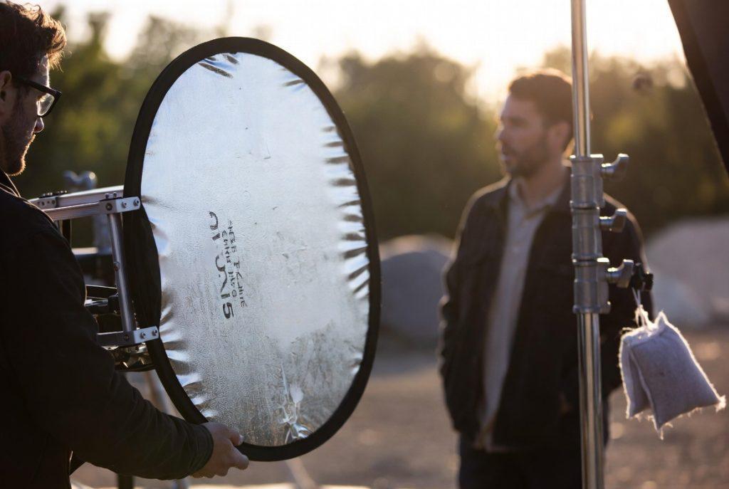 Filmmaker on outdoor set holding 5-in-1 reflector next to actor during golden hour exterior dialogue scene. Visible C-stand, sandbag, and natural sunlight. Documentary-style behind-the-scenes photo showing real production environment, not posed tutorial. 35mm depth of field, slight grain.