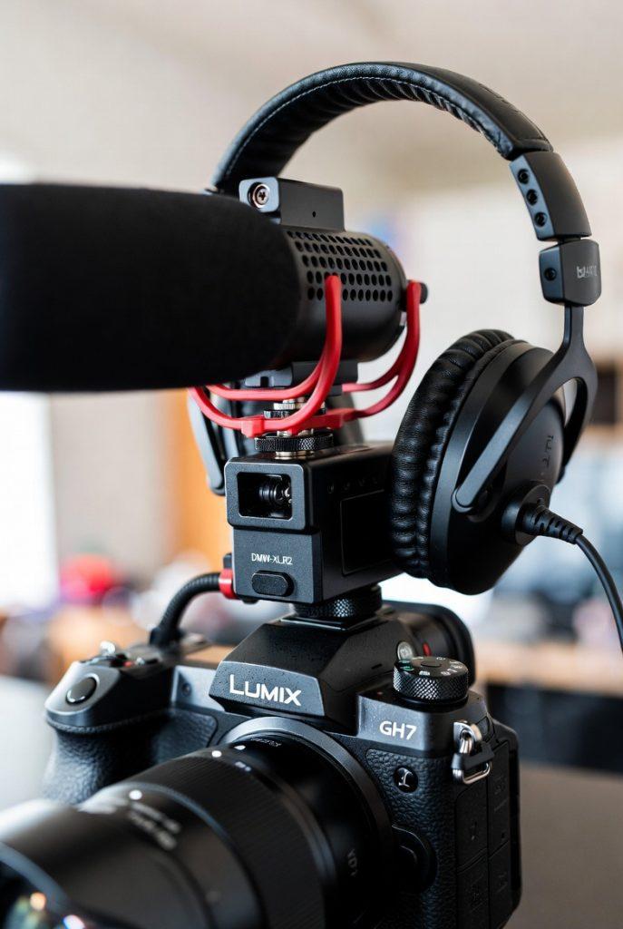 Panasonic GH7 camera with DMW-XLR2 audio adapter mounted on top, professional shotgun microphone attached, headphones connected, documentary/run-and-gun setup, emphasis on audio capabilities, practical production environment