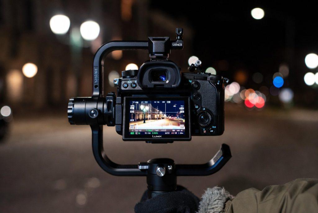 Create a realistic, cinematic photograph of a Panasonic Lumix S5 II full-frame mirrorless camera mounted on a professional gimbal during a night exterior shoot on a city street. The scene is set at night with practical street lights glowing in the background, creating moody bokeh and realistic lighting. The camera's fully articulating flip-out LCD screen is clearly visible, displaying an active V-Log waveform monitor with zebra patterns and exposure data. A camera operator wearing thick winter gloves is holding and operating the gimbal, dressed for cold weather with a jacket visible. The overall atmosphere is professional film production at night — cool color tones, subtle lens flares from the street lamps, shallow depth of field, high detail, photorealistic, 8k quality, cinematic lighting. Style: photorealistic, documentary-style film set photography, natural night lighting, sharp focus on the camera and screen, atmospheric background.