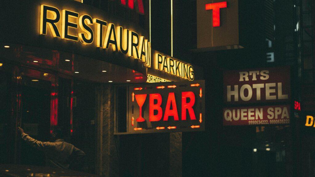 Travel filmmaker capturing nightlife in low-light conditions with neon signs and street performers in the background.