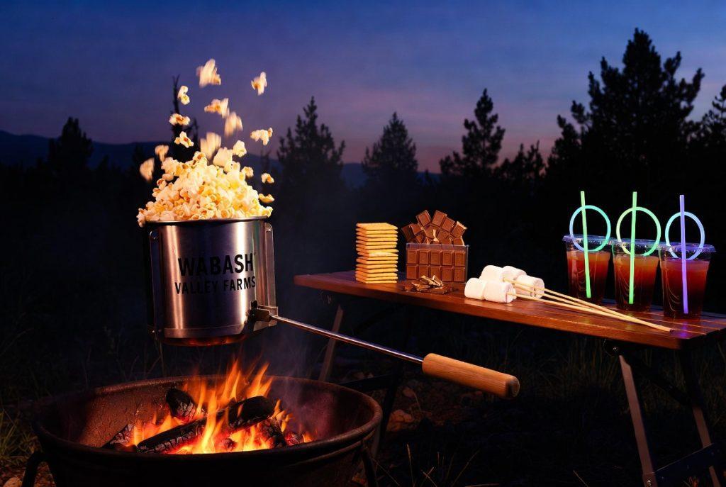 Campsite movie night snack spread — campfire popcorn popper, s'mores kit, and glow stick drinks for kids at dusk.