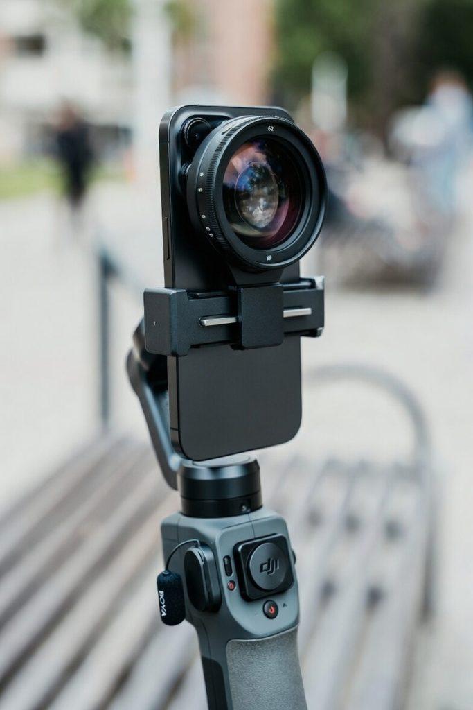 A hyper-realistic, close-up product shot focused on a modern flagship smartphone (like iPhone 17 Pro) mounted on a small, pocket-sized, dark grey stabilizer grip (such as a DJI Osmo Mobile). The phone has a compact Variable ND filter screwed onto the lens. A tiny, nearly invisible wire for a Boya lav mic is clipped neatly to the grip, disappearing into a casual jacket pocket (suggesting a wireless receiver is hidden inside). The background is a soft-focus urban park bench. The entire setup is minimal, black-on-black, and low-profile. The depth of field is very shallow, making the minimal rig the star. Cinematic, high-detail texture.