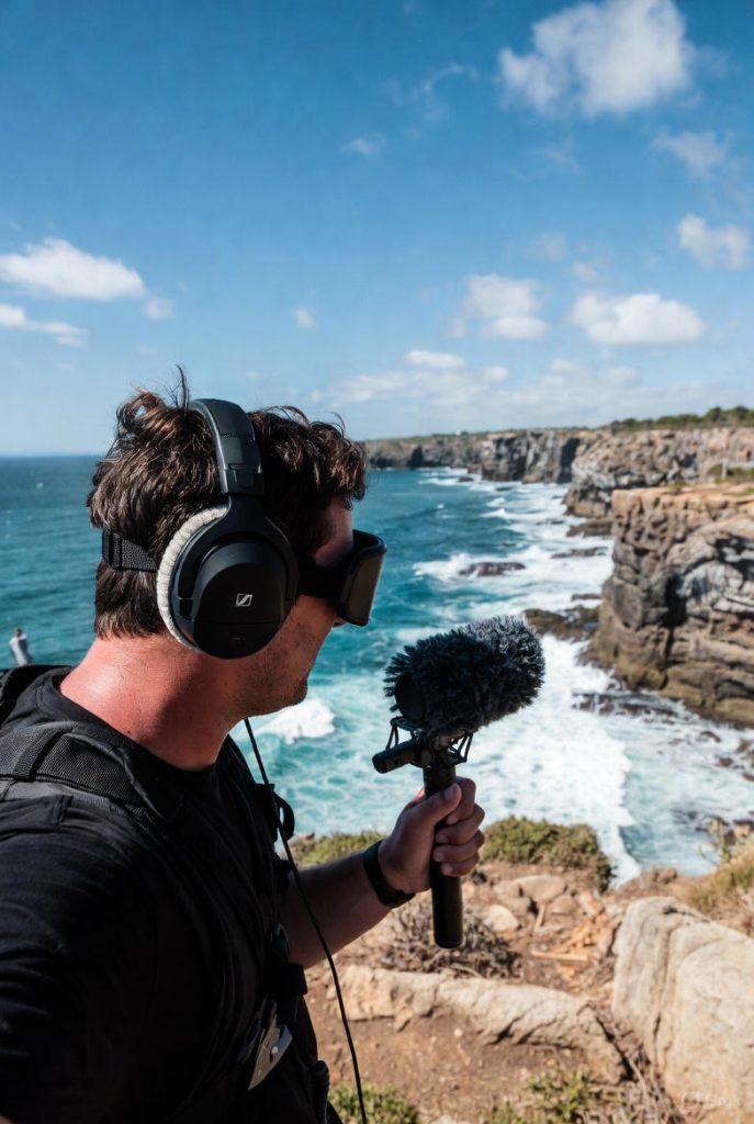 Travel vlogger wearing a wide-brimmed hat, sunglasses, and backpack, standing on a cliffside overlooking dramatic crashing ocean waves and rugged coastline under a partly cloudy sky, holding a handheld ambisonic microphone on a short boom pole with furry windshield to record immersive 360-degree spatial audio.