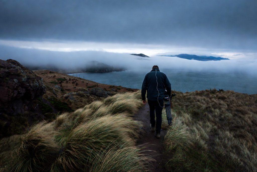 Budget wireless lav mics Trent Peek, from behind, walking the coastal trail at low light. Lav wire just visible at the collar. Tripod or monopod over one shoulder. Wind-flattened grass in the foreground, fog or overcast sky. This is the hero image. It proves the location claim immediately.