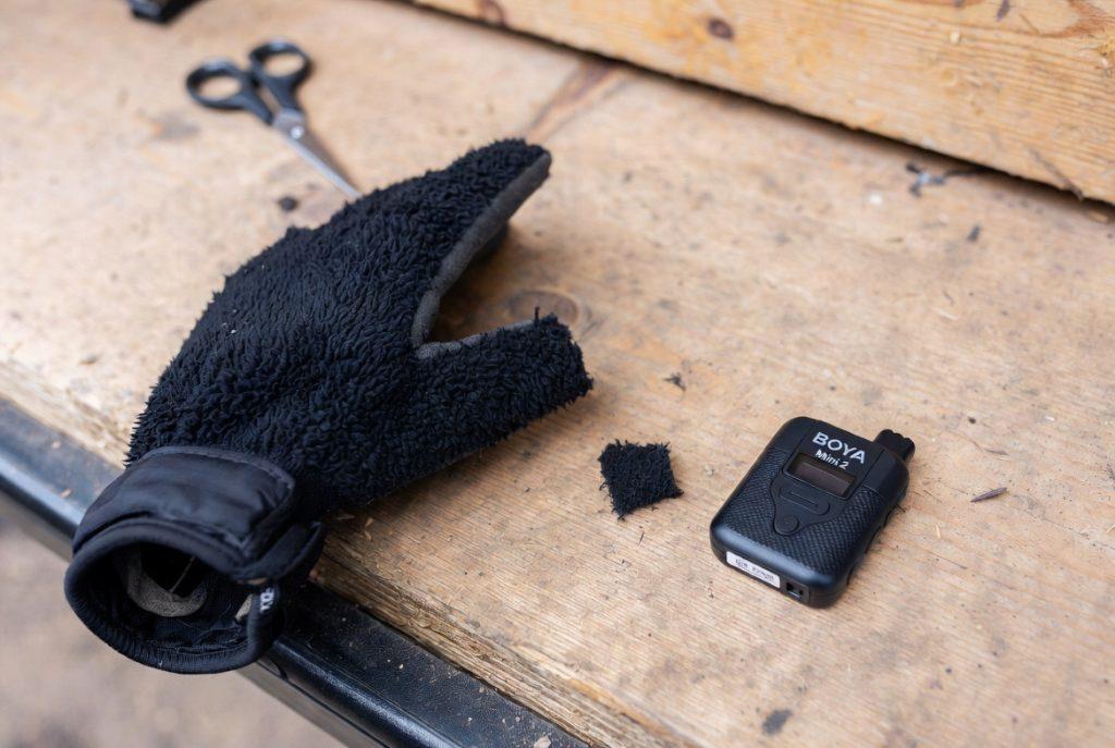 Close-up, slightly messy. A cheap winter glove on a wooden surface or tailgate, one finger half-cut, the BOYA Mini 2 next to it. Maybe a small scissors in frame. Natural light, not a product shot. The imperfection is the point.
