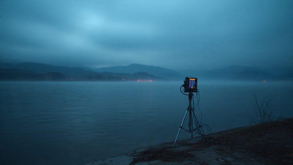 A C-stand in the foreground with an ambisonic recorder mounted on it, alone in frame, at the edge of a lake at pre-dawn. The recorder's LED is visible but you can't tell if it's recording. Slightly underexposed. The composition should feel uncertain.