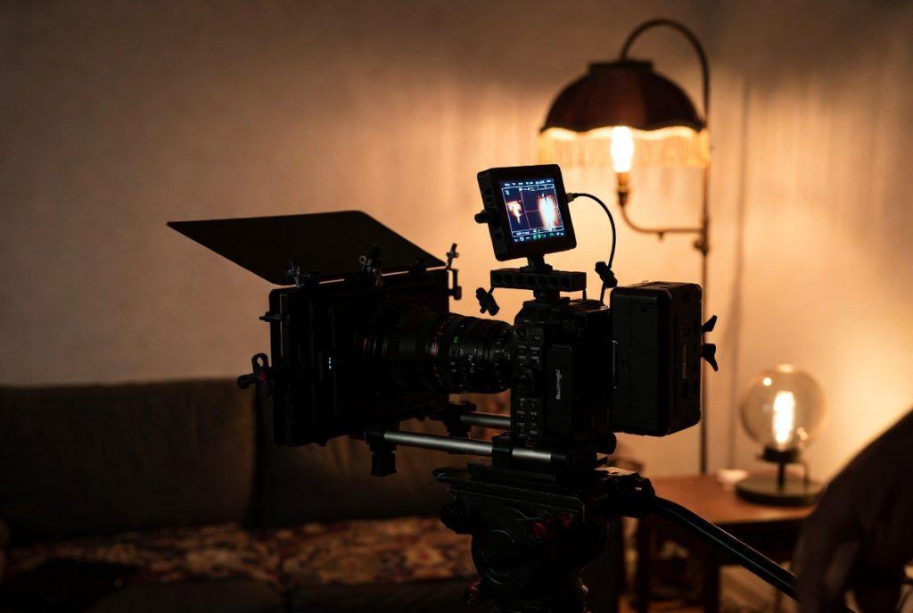 Behind-the-scenes photo of a Blackmagic camera on a tripod in a small apartment living room, with practical lighting from vintage lamps visible in the background. Moody, textured shadows. Indie film set aesthetic.