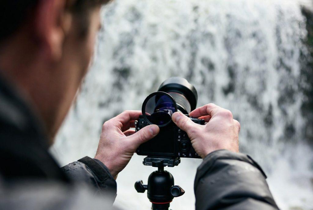 Over-shoulder shot: hands screwing an ND filter onto a lens, camera already on a travel tripod in front of a waterfall. Water visible and moving in background. Slightly wet jacket sleeves. Real location, no posing.